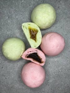 Pandangan atas pau mini berwarna hijau pastel dan merah jambu, dua daripadanya dibelah dua memperlihatkan inti kaya dan inti kacang merah, tersusun di atas permukaan kelabu.