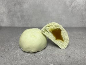 Pau mini berwarna hijau pastel dengan inti kaya yang pekat, ditampilkan satu dibelah dua di sebelah satu biji pau penuh.